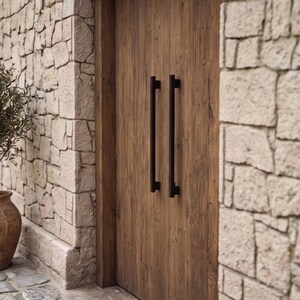 May include: A set of double wooden doors featuring a natural brown wood grain finish and two long, vertical, matte black metal pull handles. The doors are set within a frame surrounded by light-colored, irregular-shaped stone masonry walls.