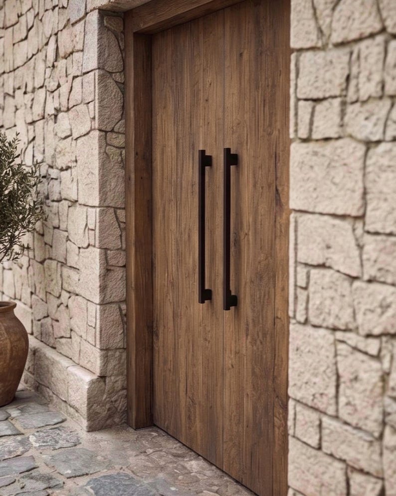 May include: A set of double wooden doors featuring a natural brown wood grain finish and two long, vertical, matte black metal pull handles. The doors are set within a frame surrounded by light-colored, irregular-shaped stone masonry walls.