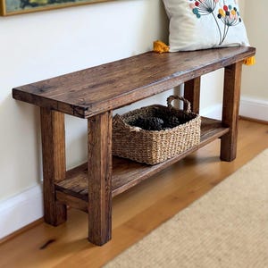 Può includere: Panca rustica in legno scuro con ripiano inferiore e cesto in vimini pieno di pigne. La panca ha un piano rettangolare. Un cuscino decorativo poggia sulla panca. La panca è lunga circa 122 cm.