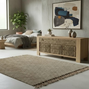 May include: A neutral-toned bedroom scene featuring a woven rug with fringe, a wooden dresser with textured drawers, and a bed with neutral bedding. An abstract painting hangs on the wall, and a potted plant adds a touch of greenery.