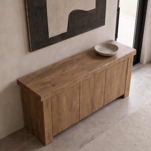 Peut inclure: Une console rectangulaire en bois avec une finition &agrave; grain naturel et des panneaux verticaux sur les portes avant. Un bol circulaire peu profond, gris et mouchet&eacute;, est pos&eacute; sur le dessus de la table, plac&eacute;e contre un mur avec une &oelig;uvre.