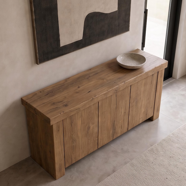 Peut inclure: Une console rectangulaire en bois avec une finition &agrave; grain naturel et des panneaux verticaux sur les portes avant. Un bol circulaire peu profond, gris et mouchet&eacute;, est pos&eacute; sur le dessus de la table, plac&eacute;e contre un mur avec une &oelig;uvre.