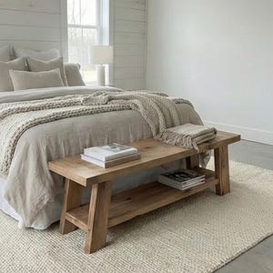 May include: A light-colored bedroom scene featuring a wooden bench with a lower shelf, holding books and magazines. The bench is positioned at the foot of a bed with neutral-toned bedding and a textured throw. A cream-colored rug and a window with a white frame complete the room.