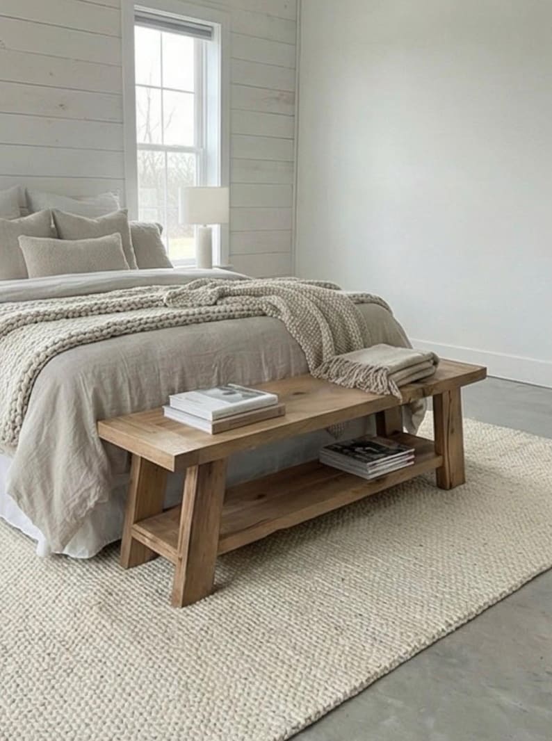 May include: A light-colored bedroom scene featuring a wooden bench with a lower shelf, holding books and magazines. The bench is positioned at the foot of a bed with neutral-toned bedding and a textured throw. A cream-colored rug and a window with a white frame complete the room.