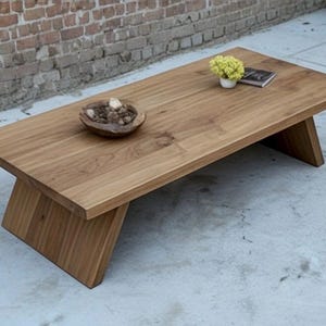 Puede incluir: Una mesa de centro rectangular de madera con acabado natural. La mesa tiene patas anguladas y una superficie lisa. Un cuenco peque&ntilde;o con objetos decorativos, una peque&ntilde;a planta en maceta y un libro est&aacute;n sobre la mesa. La mesa mide unos 152 cm de largo.