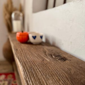 May include: A rustic wooden shelf displays a small orange pumpkin-shaped object and a white cup with blue heart designs. The shelf has a natural wood grain and is set against a white wall. A candle in a glass cloche is in the background.