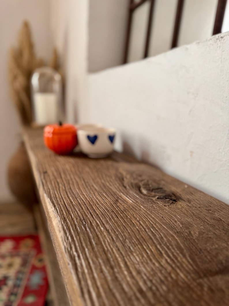 May include: A rustic wooden shelf displays a small orange pumpkin-shaped object and a white cup with blue heart designs. The shelf has a natural wood grain and is set against a white wall. A candle in a glass cloche is in the background.