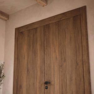May include: A set of double doors made of medium-brown wood with a visible vertical grain pattern. The doors feature a rectangular frame and a matte black horizontal handle with a matching keyhole plate positioned in the center of the doors.