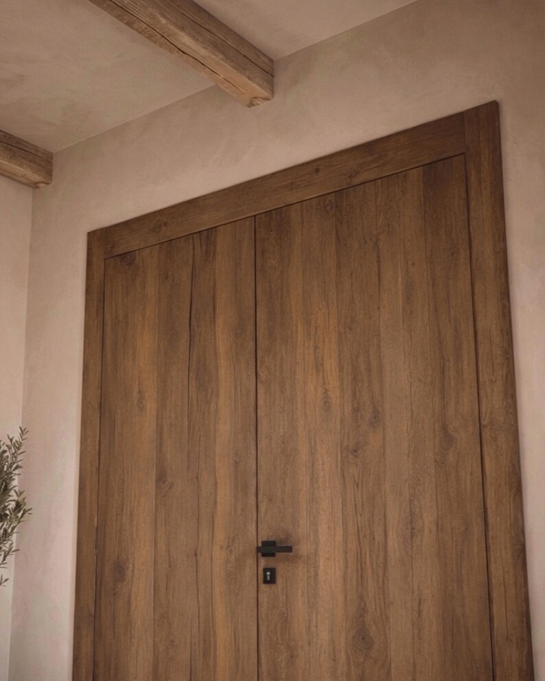 May include: A set of double doors made of medium-brown wood with a visible vertical grain pattern. The doors feature a rectangular frame and a matte black horizontal handle with a matching keyhole plate positioned in the center of the doors.