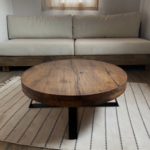 K&ouml;nnte beinhalten: Ein runder Holz-Couchtisch mit einem schwarzen Metallgestell. Die Tischplatte ist aus recyceltem Holz mit einer nat&uuml;rlichen Oberfl&auml;che. Der Tisch steht auf einem wei&szlig;en und schwarzen Teppich.
