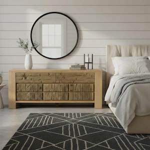May include: A bedroom scene featuring a light wood dresser with six drawers, a large round black mirror, and a gray geometric patterned rug. A bed with white bedding and a neutral headboard is on the right.