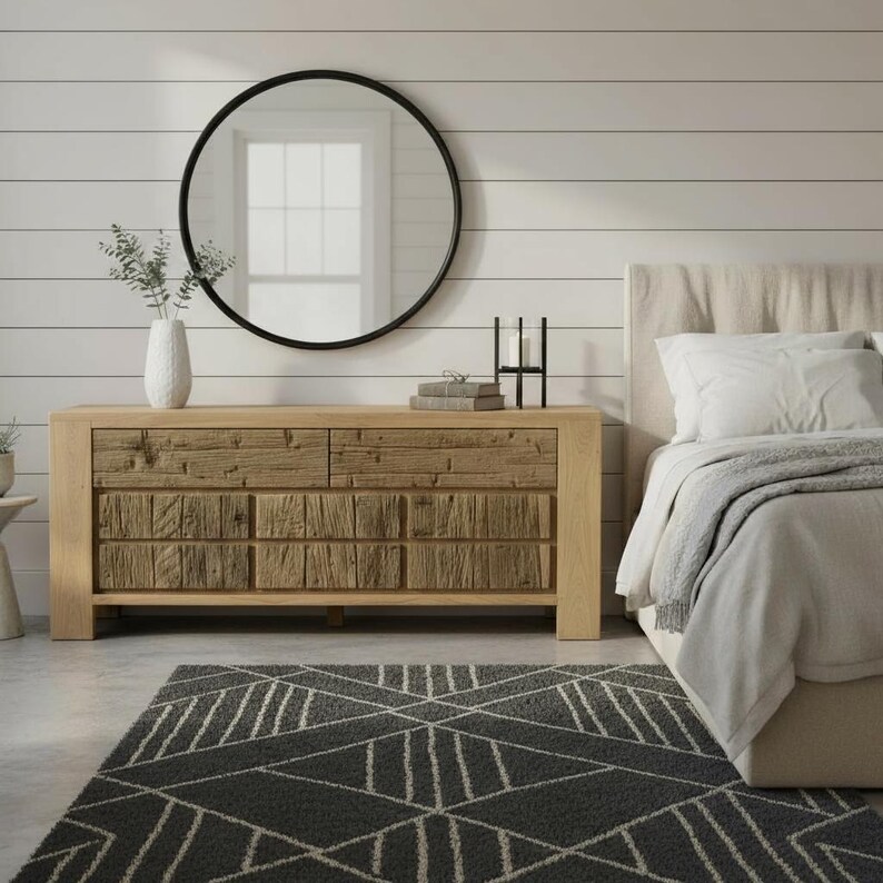 May include: A bedroom scene featuring a light wood dresser with six drawers, a large round black mirror, and a gray geometric patterned rug. A bed with white bedding and a neutral headboard is on the right.
