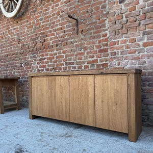 Peut inclure: Un buffet rectangulaire en bois dot&eacute; de quatre portes verticales avec une finition en grain de bois brun clair naturel. Le meuble pr&eacute;sente un plateau &eacute;pais et une structure robuste, offrant un design &eacute;pur&eacute; sans aucune quincaillerie visible.