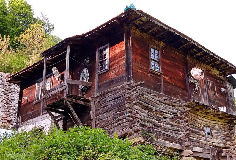 May include: A weathered, two-story wooden house with a rustic appearance. The exterior is constructed from dark brown logs and planks, featuring multiple windows, a small balcony, and a satellite dish. Green foliage surrounds the base of the structure.