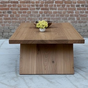 Puede incluir: Mesa de centro cuadrada de madera maciza con acabado natural. La mesa tiene una tapa gruesa y una base de pedestal. Un peque&ntilde;o cuenco de madera y una maceta blanca con flores amarillas encima.