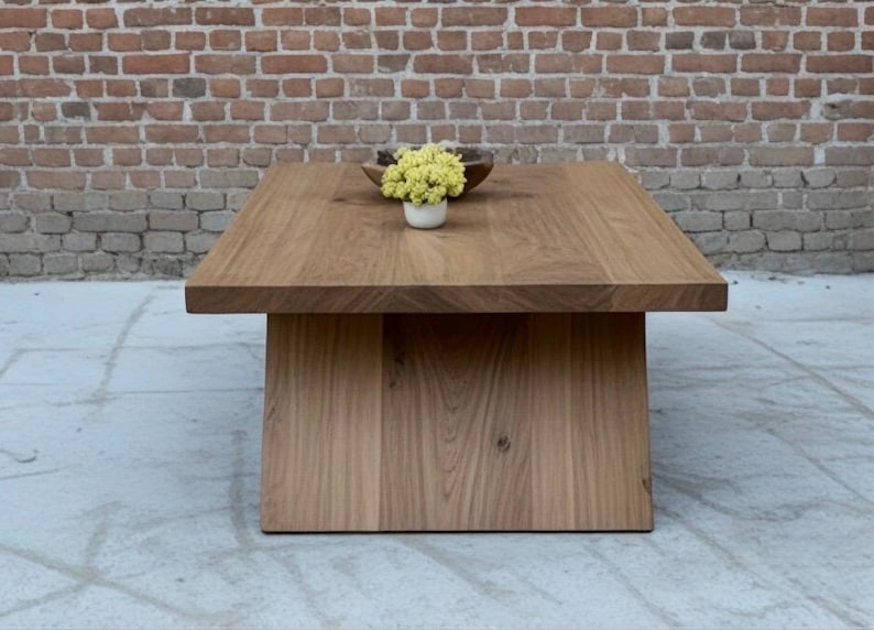 Puede incluir: Mesa de centro cuadrada de madera maciza con acabado natural. La mesa tiene una tapa gruesa y una base de pedestal. Un peque&ntilde;o cuenco de madera y una maceta blanca con flores amarillas encima.