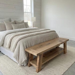May include: A bedroom scene featuring a bed with beige bedding and a chunky knit throw. A natural wood bench sits at the foot of the bed, and a neutral rug is on the floor. The walls are white with a shiplap accent.