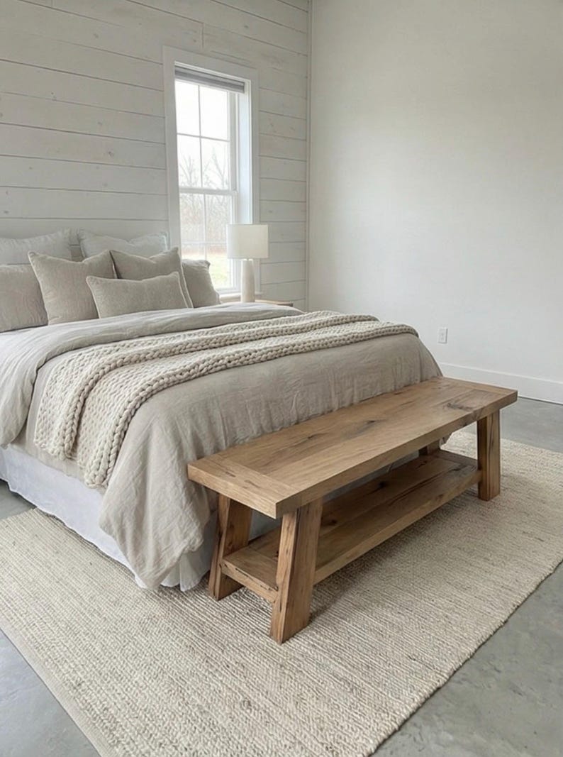 May include: A bedroom scene featuring a bed with beige bedding and a chunky knit throw. A natural wood bench sits at the foot of the bed, and a neutral rug is on the floor. The walls are white with a shiplap accent.