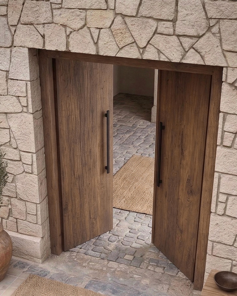 May include: A set of double doors made of medium-brown wood with a visible grain pattern. Each door features a long, vertical, matte black metal handle. The doors are set within a wall constructed of light-colored, irregular-shaped stone blocks.