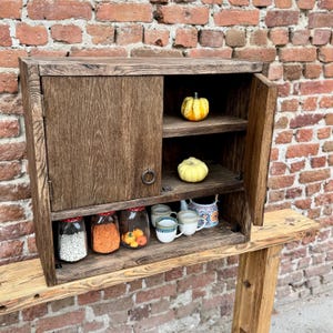 Puede incluir: Un armario de madera con una sola puerta y dos estantes. El armario est&aacute; lleno de varios art&iacute;culos, incluyendo frascos de comida, tazas y una tetera. El armario est&aacute; sentado en una encimera de madera.