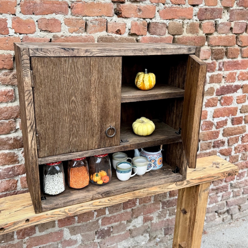 Puede incluir: Un armario de madera con una sola puerta y dos estantes. El armario est&aacute; lleno de varios art&iacute;culos, incluyendo frascos de comida, tazas y una tetera. El armario est&aacute; sentado en una encimera de madera.