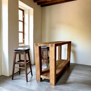 Peut inclure: Une console en bois massif avec une &eacute;tag&egrave;re inf&eacute;rieure et un tabouret de bar assorti avec une assise en cuir noir. La table est en bois brun clair et de forme rectangulaire. Le tabouret est brun fonc&eacute;.