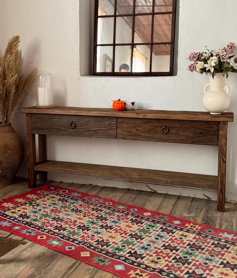 May include: A rustic wooden console table with two drawers and a lower shelf. The table is made of dark-stained wood and has metal ring pulls. A colorful patterned rug lies on the floor in front of the table. A vase of flowers and decorative items are on the table.