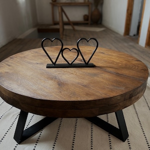 K&ouml;nnte beinhalten: Ein runder Holz-Couchtisch mit einem schwarzen Metallgestell. Der Tisch hat eine nat&uuml;rliche Holzoptik und ist mit einer herzf&ouml;rmigen Metallsculptur verziert.