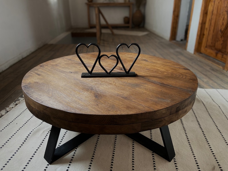 K&ouml;nnte beinhalten: Ein runder Holz-Couchtisch mit einem schwarzen Metallgestell. Der Tisch hat eine nat&uuml;rliche Holzoptik und ist mit einer herzf&ouml;rmigen Metallsculptur verziert.