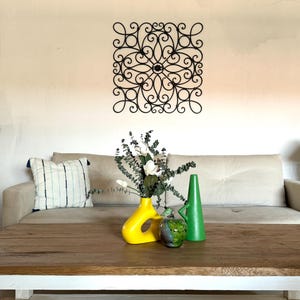 May include: A white sofa with a decorative black metal wall art piece above it. A wooden coffee table with a yellow and green ceramic vase with white flowers sits in front of the sofa.