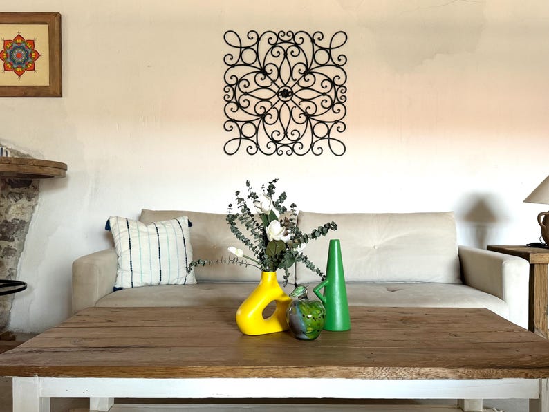 May include: A white sofa with a decorative black metal wall art piece above it. A wooden coffee table with a yellow and green ceramic vase with white flowers sits in front of the sofa.