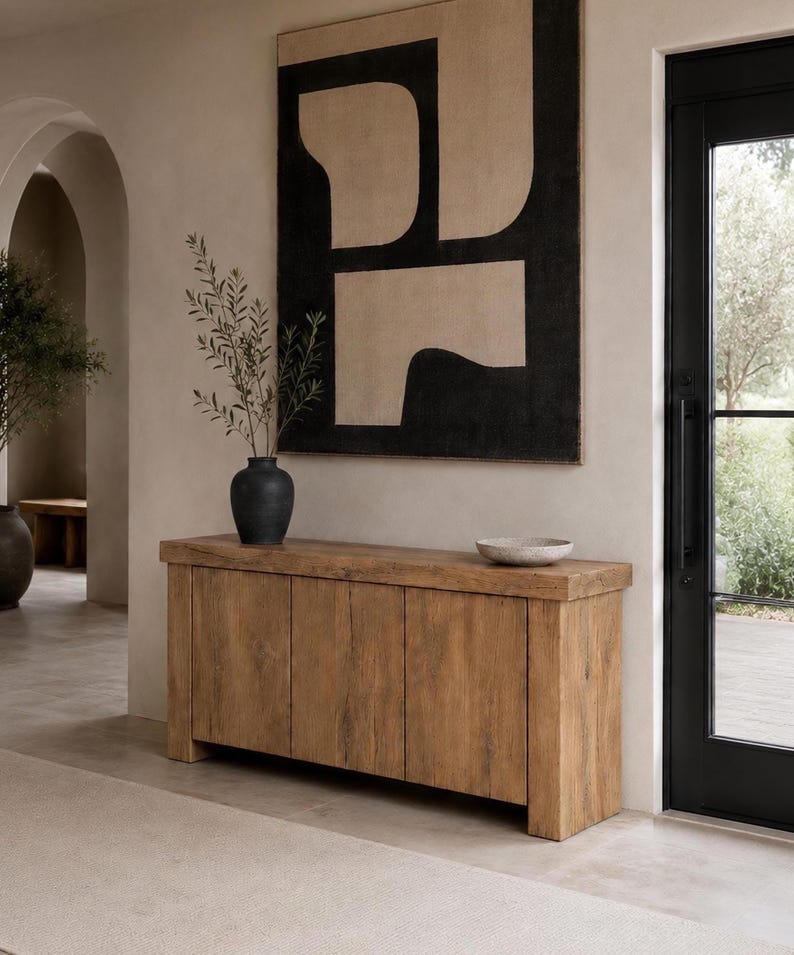 Peut inclure: Un buffet rectangulaire en bois dot&eacute; de quatre portes verticales avec une finition au grain naturel. Sur le dessus se trouvent un vase noir mat contenant des branches vertes et un bol en pierre mouchet&eacute;. Au-dessus est accroch&eacute;e une grande &oelig;uvre d'art abstraite noire et beige.
