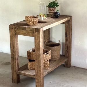 K&ouml;nnte beinhalten: Ein rustikaler Holztisch mit einem Regal, auf dem verschiedene geflochtene K&ouml;rbe und eine Glasglocke mit einer kleinen Pflanze stehen. Der Tisch hat eine nat&uuml;rliche Holzoptik und ist aus recyceltem Holz gefertigt.