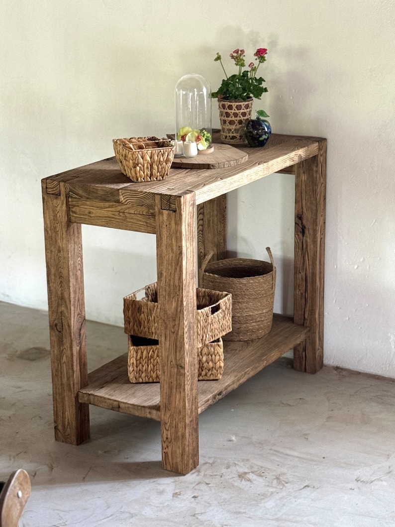 K&ouml;nnte beinhalten: Ein rustikaler Holztisch mit einem Regal, auf dem verschiedene geflochtene K&ouml;rbe und eine Glasglocke mit einer kleinen Pflanze stehen. Der Tisch hat eine nat&uuml;rliche Holzoptik und ist aus recyceltem Holz gefertigt.
