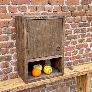 Puede incluir: Un armario de madera marr&oacute;n oscuro con una sola puerta y un peque&ntilde;o estante. El armario tiene una manija de anillo de metal. El armario est&aacute; sentado en un estante de madera con tres peque&ntilde;as calabazas en el estante.