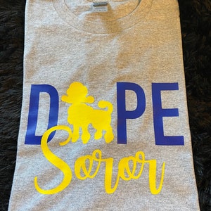 May include: Gray t-shirt with the text "Dope Soror" in blue and yellow lettering. A yellow silhouette of a poodle is in the middle of the text.