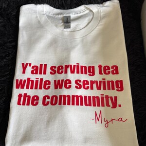 May include: White t-shirt with red text that reads "Y'all serving tea while we serving the community. -Myra"