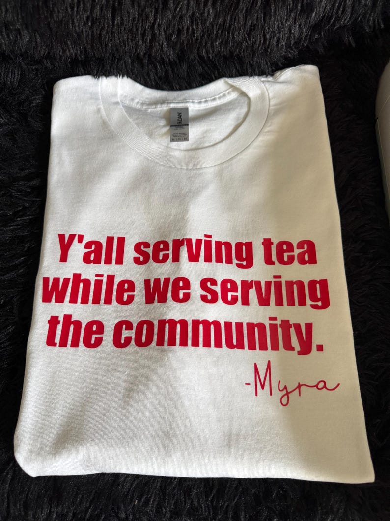 May include: White t-shirt with red text that reads "Y'all serving tea while we serving the community. -Myra"