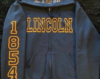 Puede incluir: Una sudadera con capucha azul marino con cremallera, con un león bordado en oro en la capucha y el texto "LINCOLN 1854" en la manga.