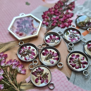 May include: A collection of silver-toned metal jewelry pendants featuring pressed pink heather flowers encased in clear resin. The set includes one hexagonal pendant and several circular pendants with small loops for attachment to chains or cords.