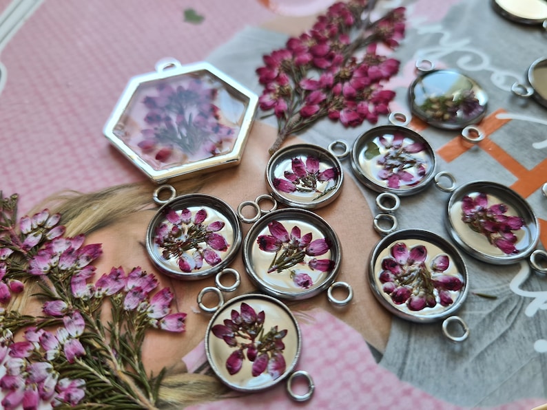May include: A collection of silver-toned metal jewelry pendants featuring pressed pink heather flowers encased in clear resin. The set includes one hexagonal pendant and several circular pendants with small loops for attachment to chains or cords.