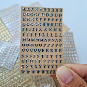 May include: A rectangular sheet of adhesive stickers featuring the English alphabet in uppercase letters. The letters are metallic silver with a dark outline, arranged in rows on a tan backing. Multiple identical sheets are visible in the background.