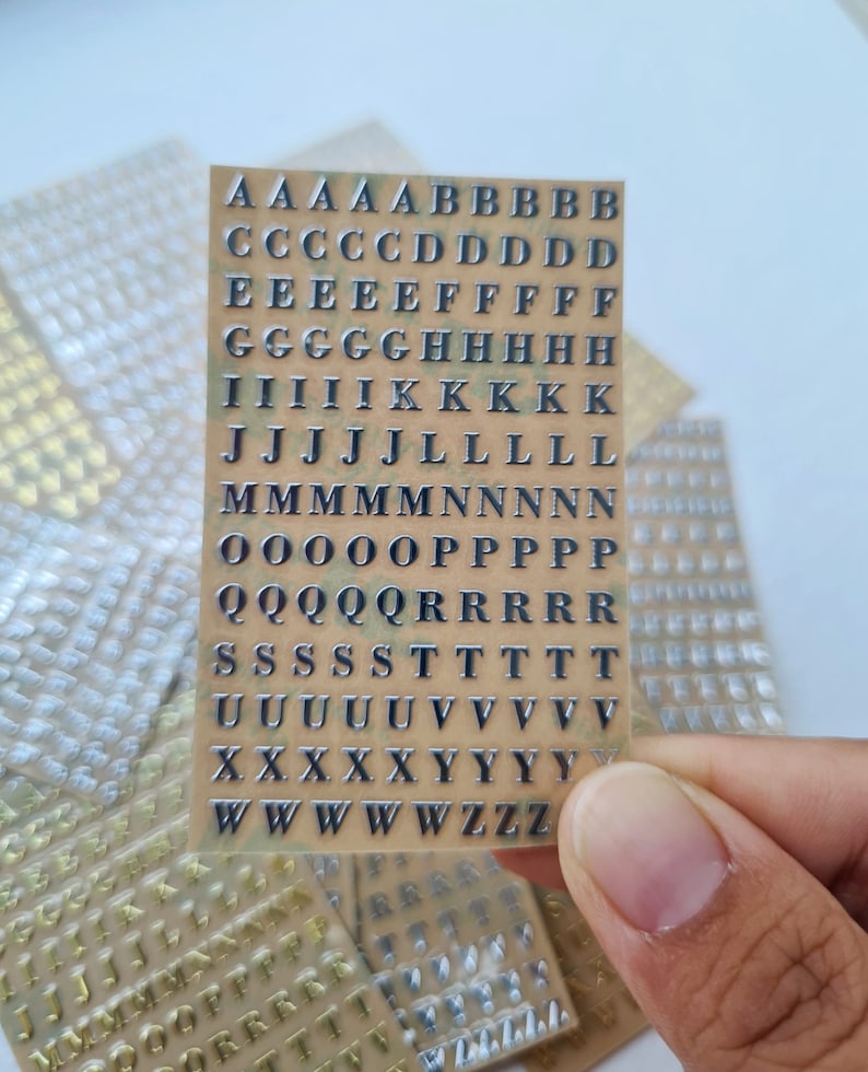 May include: A rectangular sheet of adhesive stickers featuring the English alphabet in uppercase letters. The letters are metallic silver with a dark outline, arranged in rows on a tan backing. Multiple identical sheets are visible in the background.