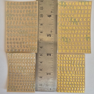 May include: Four sheets of adhesive alphabet stickers featuring uppercase and lowercase letters in silver and gold metallic finishes. A metal ruler is positioned vertically between the sheets, showing a length of approximately 14 cm.