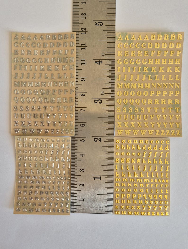 May include: Four sheets of adhesive alphabet stickers featuring uppercase and lowercase letters in silver and gold metallic finishes. A metal ruler is positioned vertically between the sheets, showing a length of approximately 14 cm.