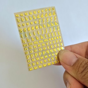 May include: A rectangular sheet of adhesive stickers featuring multiple rows of lowercase letters in a metallic gold finish. The letters are arranged in alphabetical order, with several duplicates of each character provided on the clear backing sheet.