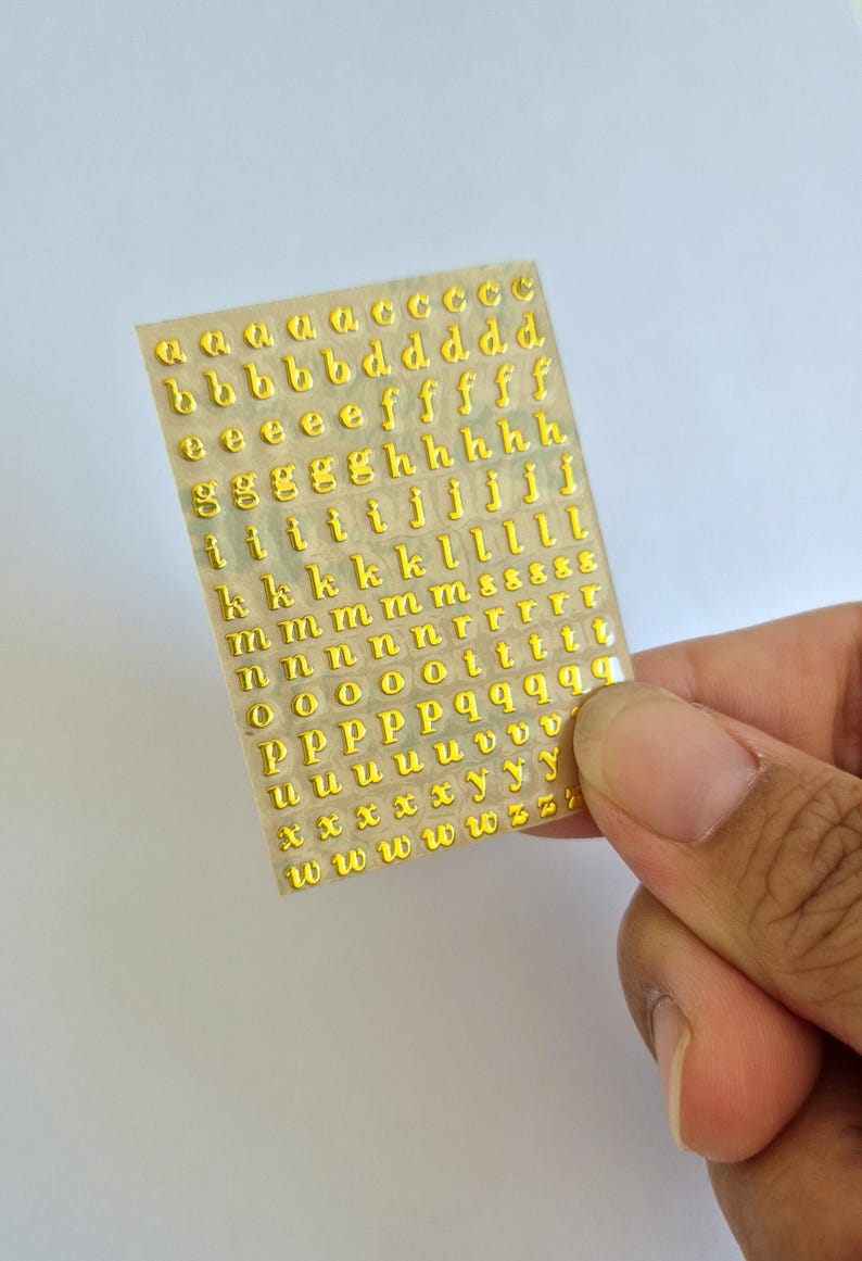 May include: A rectangular sheet of adhesive stickers featuring multiple rows of lowercase letters in a metallic gold finish. The letters are arranged in alphabetical order, with several duplicates of each character provided on the clear backing sheet.