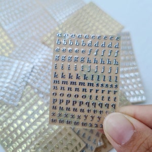 May include: A rectangular sheet of small, metallic silver adhesive lowercase alphabet stickers arranged in neat rows. The letters include multiple instances of each character, such as a, b, d, e, f, g, h, i, j, k, l, m, n, o, p, q, r, s, t, u, w, x, y, and z.