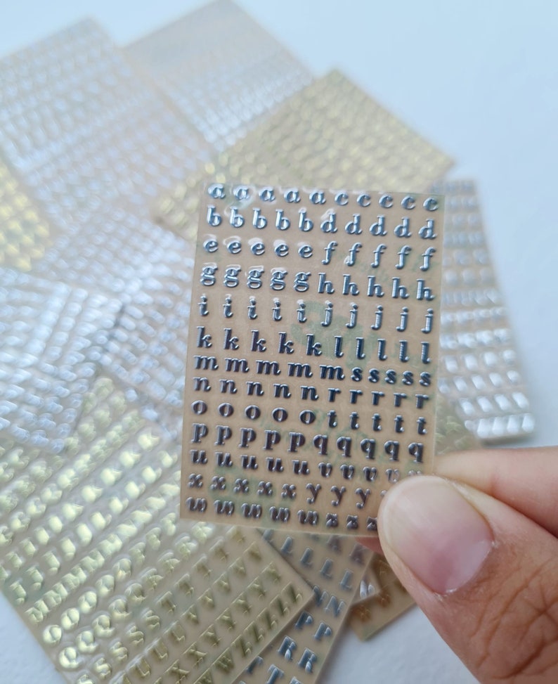 May include: A rectangular sheet of small, metallic silver adhesive lowercase alphabet stickers arranged in neat rows. The letters include multiple instances of each character, such as a, b, d, e, f, g, h, i, j, k, l, m, n, o, p, q, r, s, t, u, w, x, y, and z.