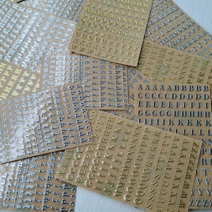 May include: Multiple rectangular sheets of adhesive stickers featuring metallic gold and silver alphabet letters in both uppercase and lowercase fonts. The stickers are arranged in organised rows and columns on clear backing sheets, showcasing various letters.